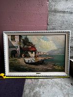 Framed oil painting showing a coastal village with buildings, boat, and figures by the water under a cloudy sky.