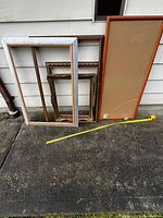 Four assorted picture frames leaning against exterior siding on pavement, includes large light brown wooden frame with beige backing, large white wooden frame, medium and small ornate gold-tone frames, with visible scratches on some.
