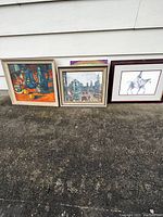 Three framed paintings leaning against exterior wall on concrete