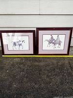 Two framed watercolor paintings showing horses and riders placed side by side on a concrete surface outdoors, frames with dark red wood and double mats.