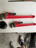 Two red Proto brand adjustable pipe wrenches (sizes 8" and 14") alongside a large black hex pipe socket on a white surface.