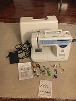Front view of sewing machine with power cord, white hard case cover behind, various small sewing machine accessories and manual spread out in front.