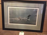 Framed Robert Bateman lithograph 'The Loons' showing two loons swimming in water, overall view of framed piece lying on wooden floor.