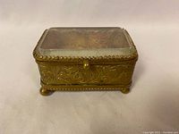Front and top angled view showing embossed brass details and patterned fabric inside the box beneath the glass lid.