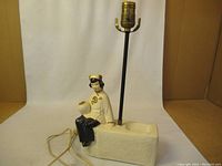 Side view of the ceramic lamp base showing the attached Asian female figurine sitting on the textured white rectangular base with black metal lamp rod and electrical cord.