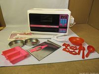 Photo showing Easy Bake Oven with baking trays, cookie cutters, spatula, mixing bowl, and measuring spoon arranged in front.