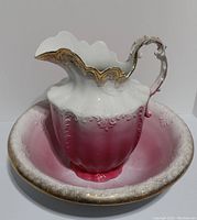 Close-up front view of the pink and white Victorian-style ironstone water pitcher sitting inside the wash basin, showing ornate gold accents on the pitcher rim and handle.