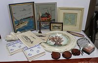 Lot of nautical-themed home decor and collectibles including needlepoints, framed art, ashtrays, trivet, tiles, and wood sculpture.