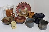 Lot laying on a table showing all items together: two wood bowls with rooster/hen motifs, ceramic hen plate, dark blue speckled enamelware pot, two small metal buckets, wooden miniature outhouse, ceramic flour shaker, and cookie cutters in plastic bag.