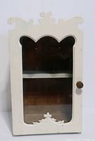 Front view of the shabby white wood wall cabinet showing the glass inset door with decorative shaped frame and scalloped trims at top and bottom.