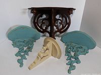 Photo showing one Victorian walnut corner wall shelf and a pair of blue painted Syroco wood wall sconce shelves arranged on a white background.