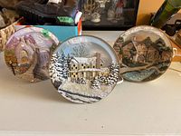 Three ceramic village scene plates arranged in a row showing detailed 3D relief work and glazed finish.