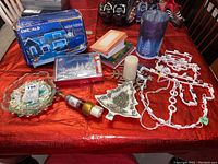 Assorted Christmas decoration items laid out on a red fabric surface, including dishes, a white candle, a ceramic tree dish, string lights, glass candle holder, small trivet, and card holder.