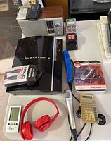 Various electronics displayed on table including PS3 console, shelf radio, MP3 player, headphones, microphones, voice recorder, chargers, and cable.