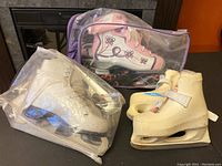 Three pairs of girls' ice skates in protective plastic bags: white Bauer, Lange Galaxy with butterfly pattern, and cream Reebok Glitter Girl 2.