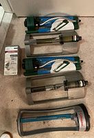 Five lawn sprinklers arranged on floor: two green Orbit/Nelson impact sprinklers, two gray and blue Gilmour oscillating sprinklers, one smaller package box of sprinkler nozzles.