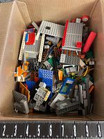 Top-down view showing assortment of Lego parts in mixed colors including gray, red, blue, orange, with plates, bricks, and specialty pieces such as windows and ladders