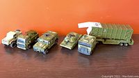Five plastic miniature army vehicle scale models including a missile launcher truck, a missile multiple rocket launcher truck, a tank, a military truck, and a beige armored jeep in camouflage colors on a brown surface against an orange background.