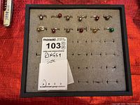 Top-down view of 12 rings mounted on gray felt ring display tray with black frame, some rings featuring red, green, and gold bell charms.