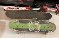 Photo shows pair of Head child skis with bindings and two skateboards stacked side by side on a floor.
