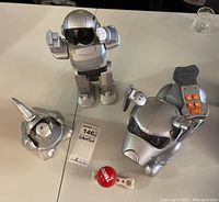 Top-down image showing three silver Tekno toy robots: a standing humanoid Robot Ditto, a Robot Dog with orange buttons and a small Robot Parrot Polly perched on its head. Also a red Tekno branded ball shaped accessory with white handle.