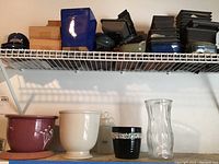 Overhead shelf holding an assortment of bonsai pots and trays in ceramic finishes in various colors and shapes, alongside a few decorative pots and a glass vase on the lower surface.