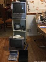 Overall view of the white rolling bookcase holding a Sylvania TV/VCR combo unit on top, a small TV with remotes on the middle shelf, and JWin portable TV on the floor with two portable DVD players. Also visible is another Sylvania TV/VCR unit on floor in front of bookcase.