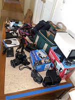 Overview photo showing multiple cameras, boxes, cases, cables, quick charger, straps and additional accessories on wooden table.