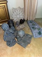 Photo of yard statues including faux stone angel child holding a leaf, little boy reading a book, stone floral wall art pieces, and ceramic gutter drainage item with frog decorations on a carpet.