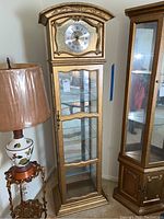Full front view of tall narrow curio cabinet with clock at top, glass doors and shelves, gold-tone finish.