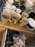 Photo showing stacked Corelle plates and bowls with beige and white coffee mugs, along with pile of dishes on countertop.