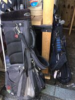 Photo showing two black golf bags with golf clubs and zippered pockets. The bags are worn and dusty.