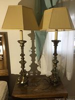 Pair of 41 inch metal table lamps with beige fabric lampshades placed on dark wooden furniture in front of white curtain.
