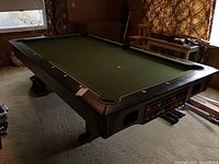 Full view of the pool table showing green felt playing surface, wood frame, corner wear, and pool balls.