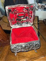 Photo of open decorative footed silver-tone keepsake box with red lining, showing floral embossed interior and exterior.