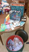Wide angle image showing the sewing and craft supplies on a round table, including storage drawers with beads, a green patterned tote bag, postal scale, candle holder, assorted notions, and a red bucket/bin with yarn and crochet needle below the table