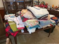 Photo of assorted stacked table linens including tablecloths, napkins, hand towels, and placemats on wooden table