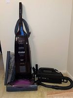 Full view of Kenmore Progressive upright vacuum in dark maroon color, purple brush base, with power cord wrapped and multiple loose vacuum attachments around it along with black Oreck XL handheld vacuum with hose.