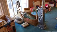 Photo shows multiple vacuum cleaners and accessories arranged on carpeted floor including Electrolux Carpet Beautifier, Bissell Sweep Easy, Electrolux Automatic G vacuum with accessories, Oreck XL with cloth bag, and box of vacuum bags.