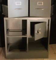 Gray metal two drawer file cabinet with an attached small metal safe. The safe door is open showing the interior. Cabinet shows light wear.