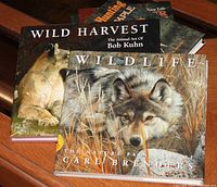 Four outdoor and wildlife art themed books stacked on a wooden surface, titles visible include 'Wild Harvest' by Bob Kuhn and 'Wildlife' by Carl Brenders.