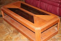 Photo showing a solid oak coffee table with two glass inserts on the top and one on the bottom shelf, placed near a red couch.