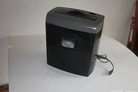 Black electric paper shredder with top-loading slot, power cord visible, placed on a white surface against a neutral background.