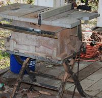 Outdoor photo of a used table saw on a metal stand with visible rust and dirt, missing the cutting blade.
