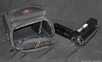 Photo showing the black Canon video camcorder with lens and flip-out screen next to the black soft carrying case with Canon logo.