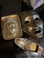 Photo of three brass decorative items: a brass plate embossed with a Buddha head, a large hollow-faced brass mask, and a decorative brass shoe with ornate engravings and cutout.