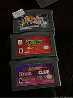 Three GameBoy Advance cartridges stacked showing front labels with game titles WWE, Frogger's Adventures, Battleship/Risk/Clue compilation.