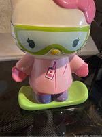 Front view showing Hello Kitty in pink ski outfit with green goggles, standing on a green snowboard.