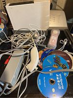 Photo showing white Nintendo Wii console, power adapter, multiple cables, white Wii Remote controller with attached Nunchuck, and several loose Wii game discs.