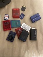 Image of various PlayStation 2 memory cards, SD cards, and adapters on a beige fabric surface.